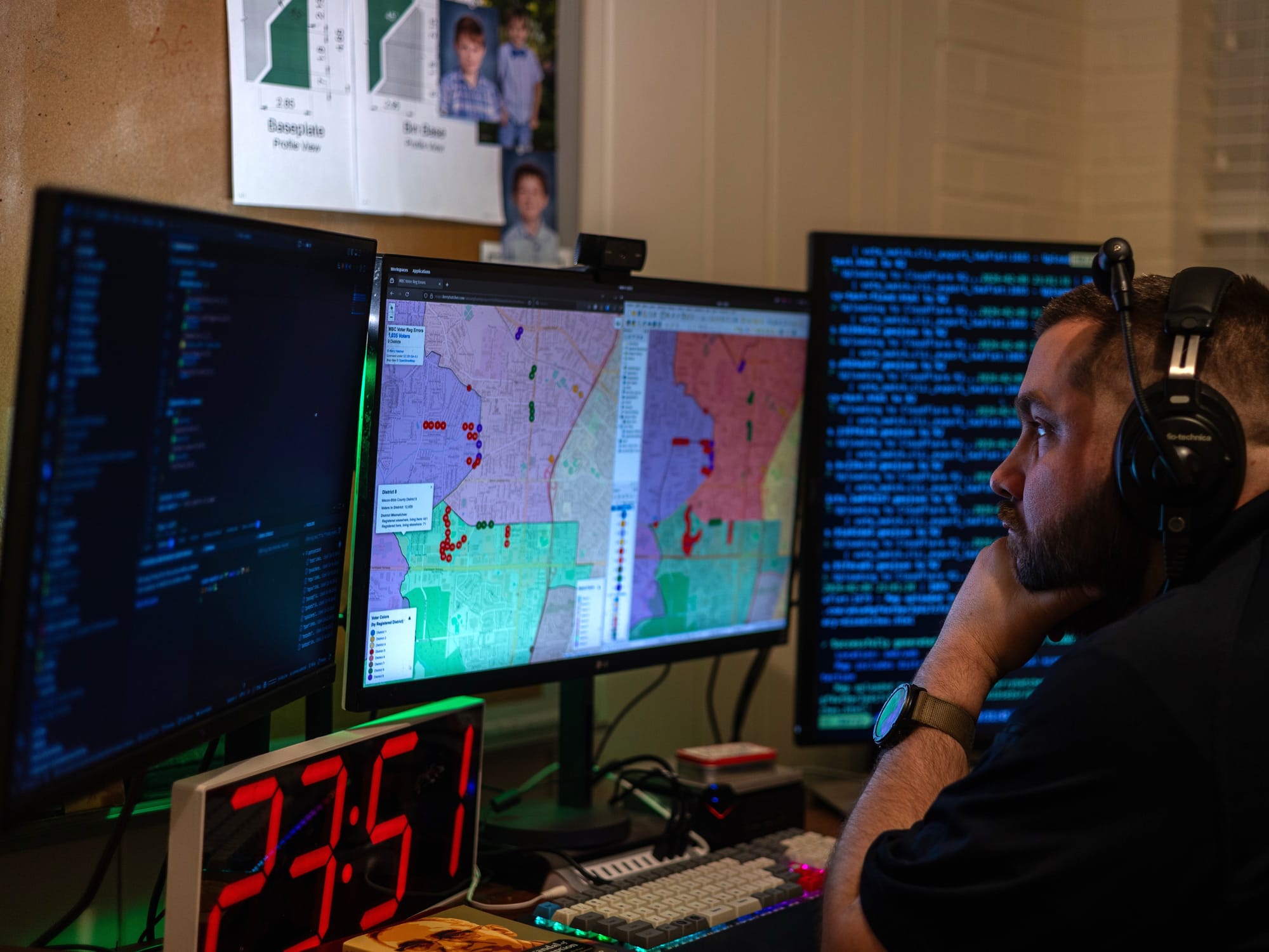 A man wearing headphones reviews a QGIS map of Macon-Bibb County voter district mismatches on a center monitor, flanked by two screens of Python code, at a desk late at night.